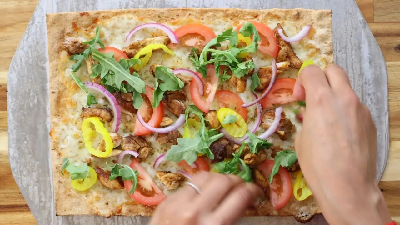 Flatbread pizza with tomatoes, chicken, red onions, banana peppers, and arugula being garnished by hand.