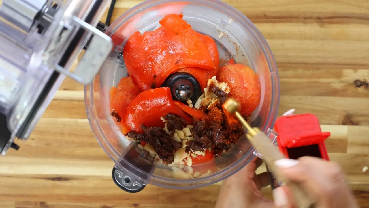 Food processor with tomatoes, nuts, and spices being prepared for a recipe on a wooden countertop.