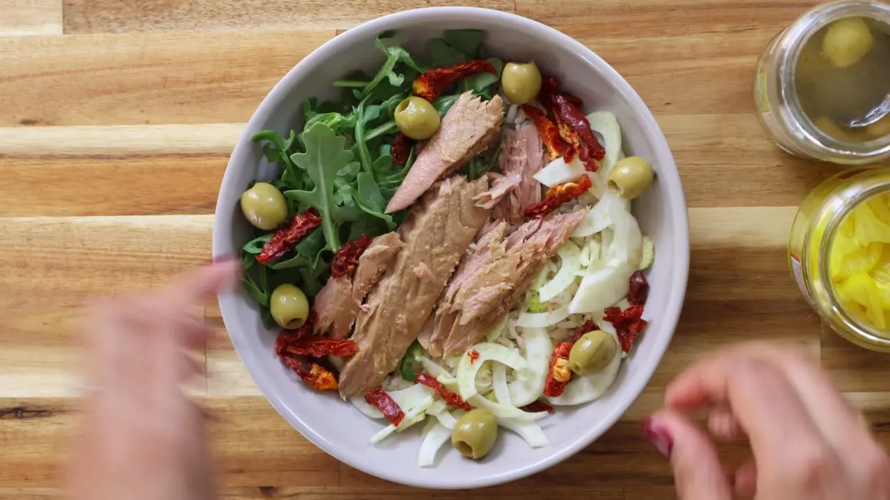Fresh salad with tuna, arugula, olives, sun-dried tomatoes, and fennel in a bowl on a wooden table.