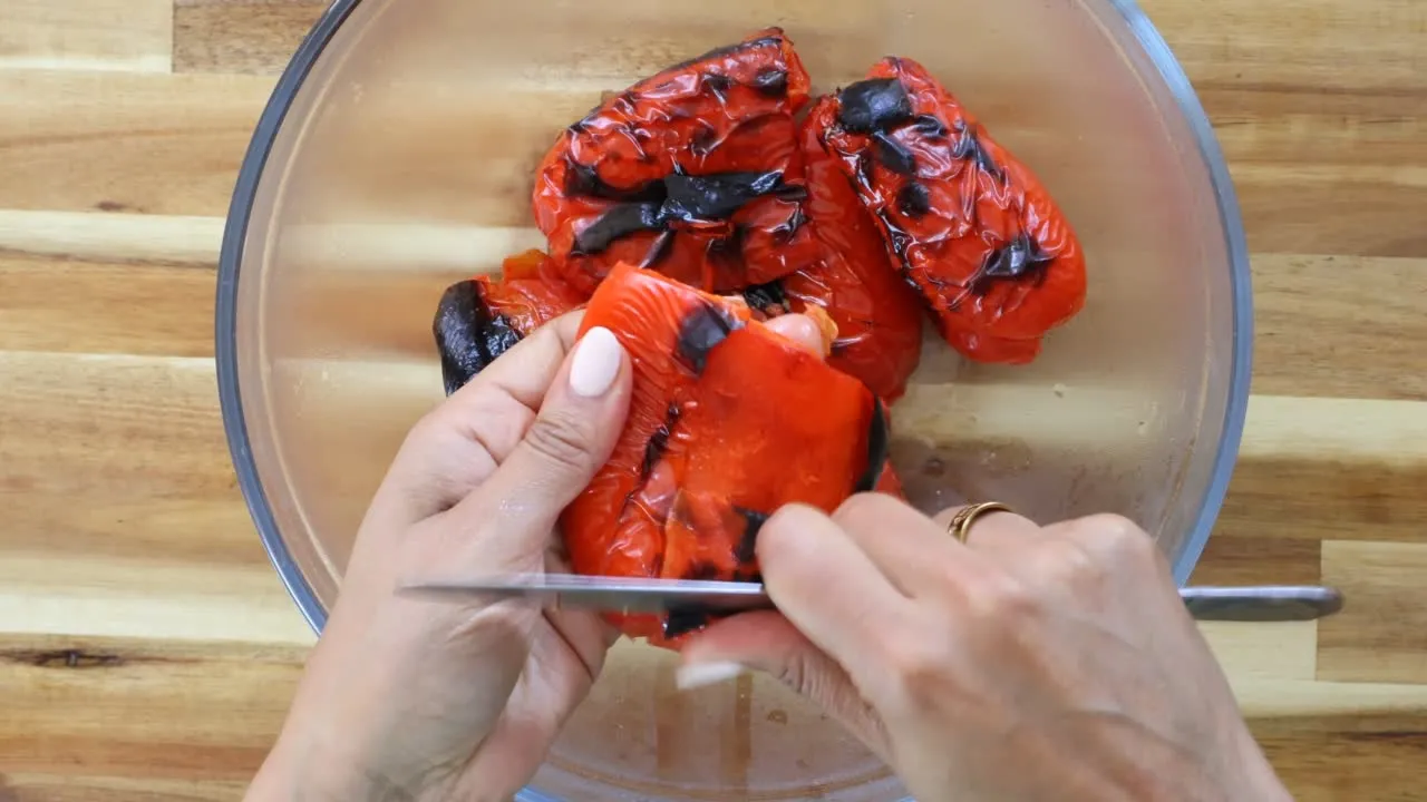 Peeling charred skin off roasted red peppers in glass bowl on wooden surface.