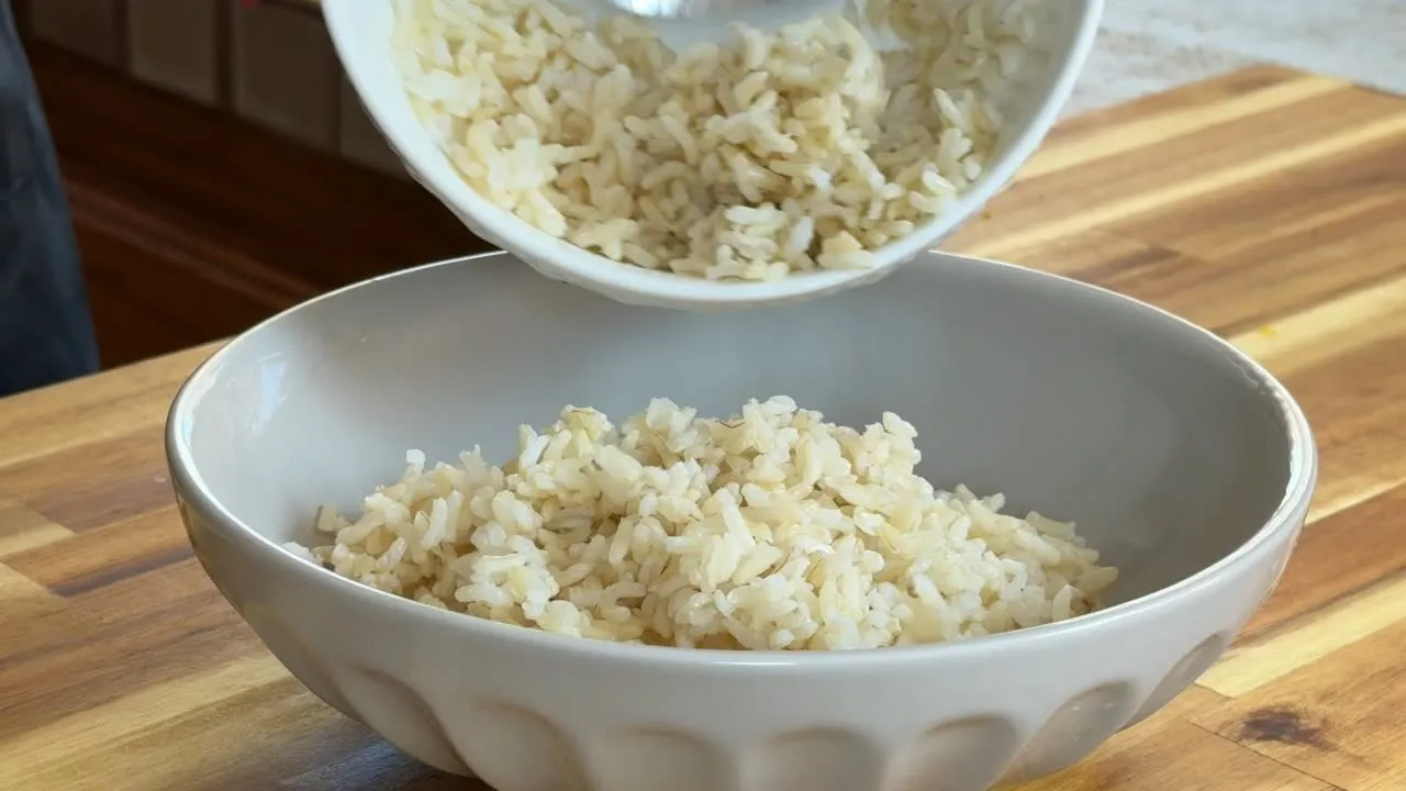 Pouring cooked brown rice into a ceramic bowl on a wooden countertop.