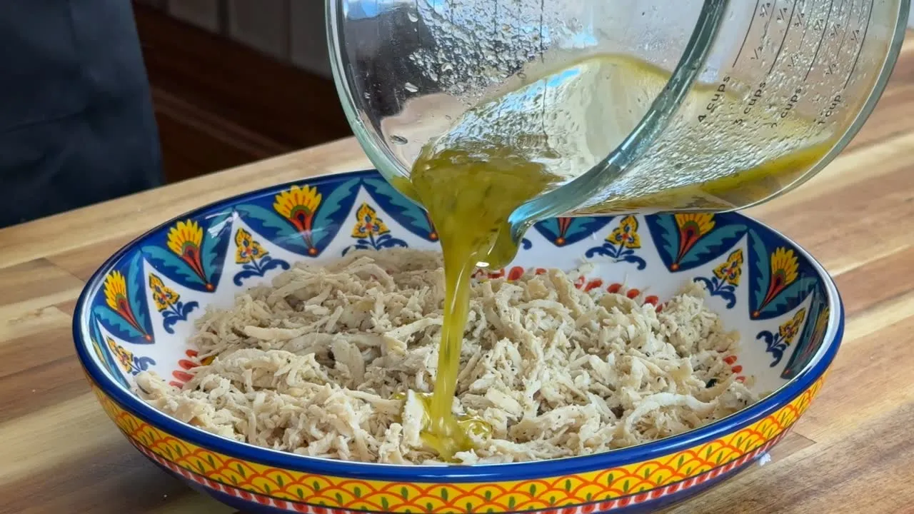 Pouring olive oil over shredded chicken in a decorative bowl on a wooden countertop.