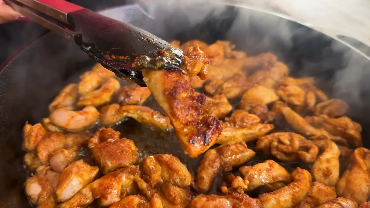 Tongs holding cooked chicken pieces above a sizzling pan, with steam rising.