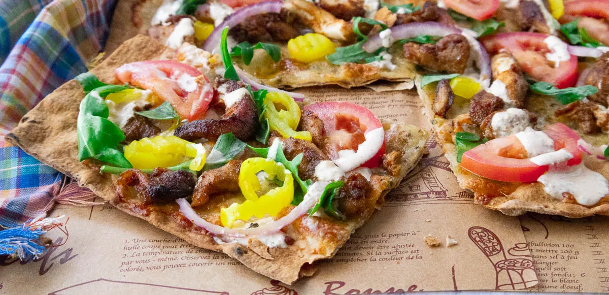 Fresh shawarma pizza topped with tomatoes, peppers, onions, and greens on printed parchment paper.