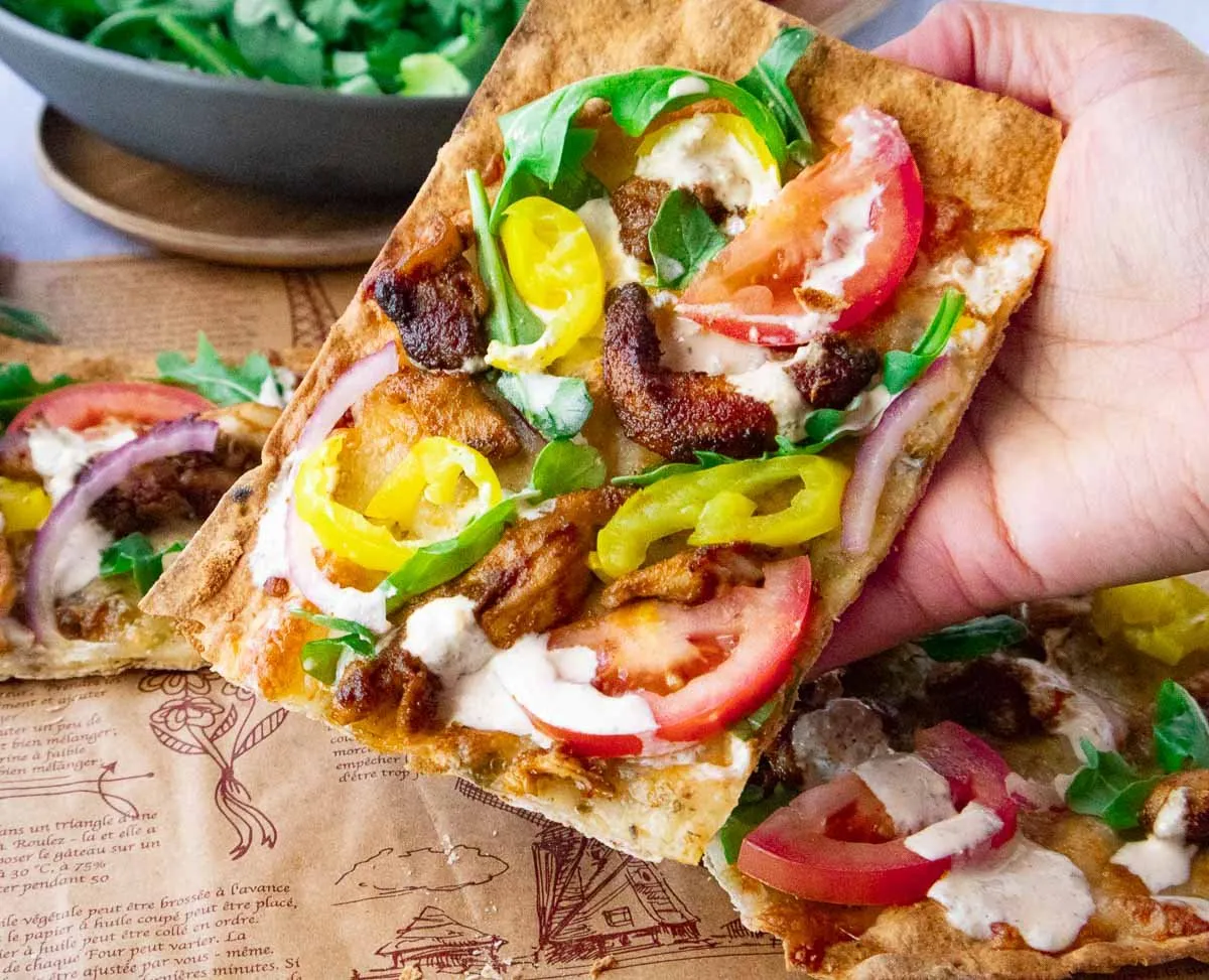 Shawarma pizza topped with tomatoes, yellow peppers, onions, and greens, held by hand over decorative paper.