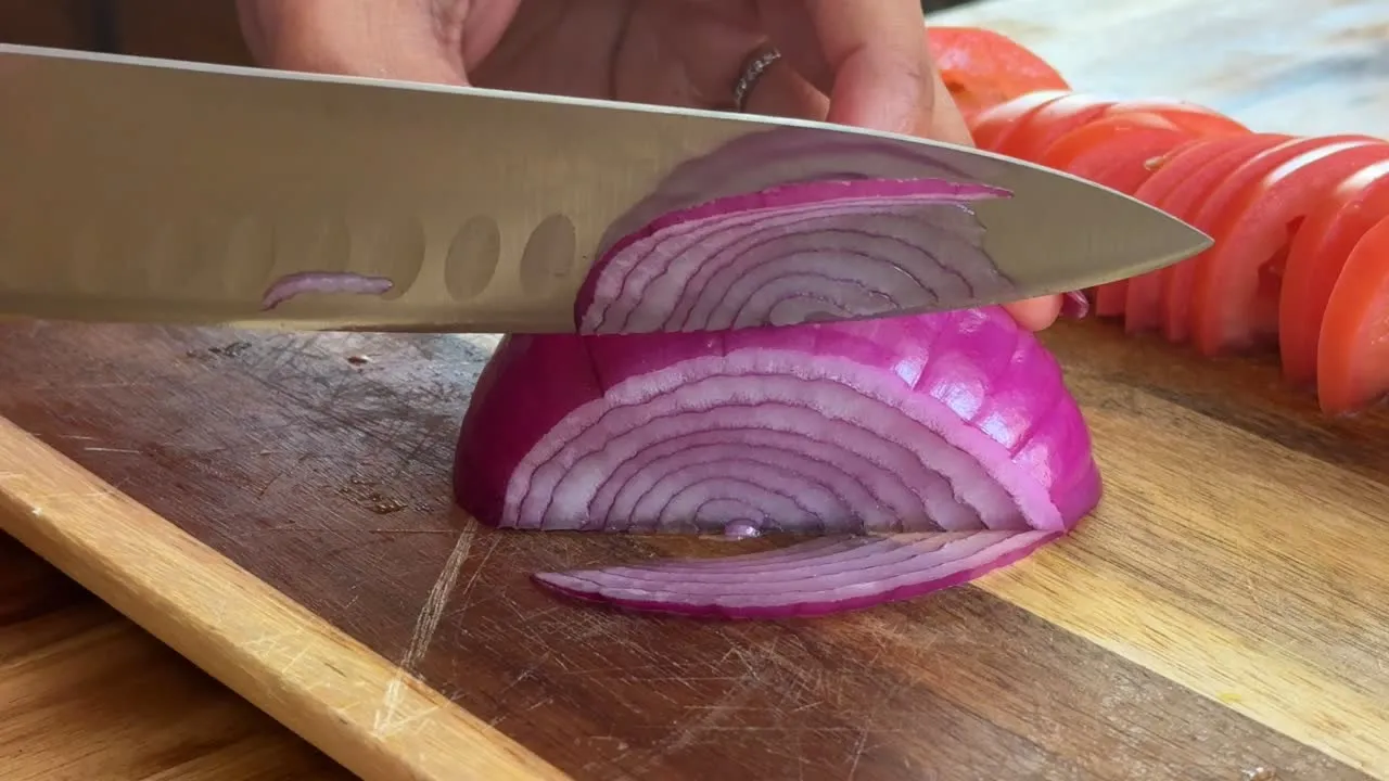 Slicing a red onion on a wooden cutting board with a knife, tomato slices in the background.