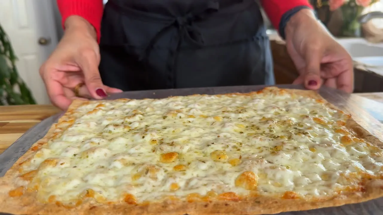 Freshly baked cheese pizza on a tray held by a person in a red sleeve and black apron.