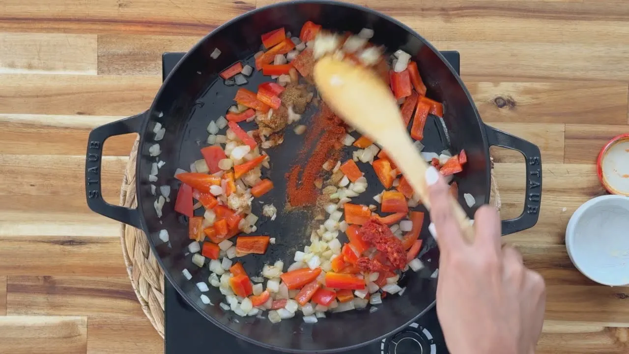 Cooking onions and red peppers in a pan with spices on a wooden surface.