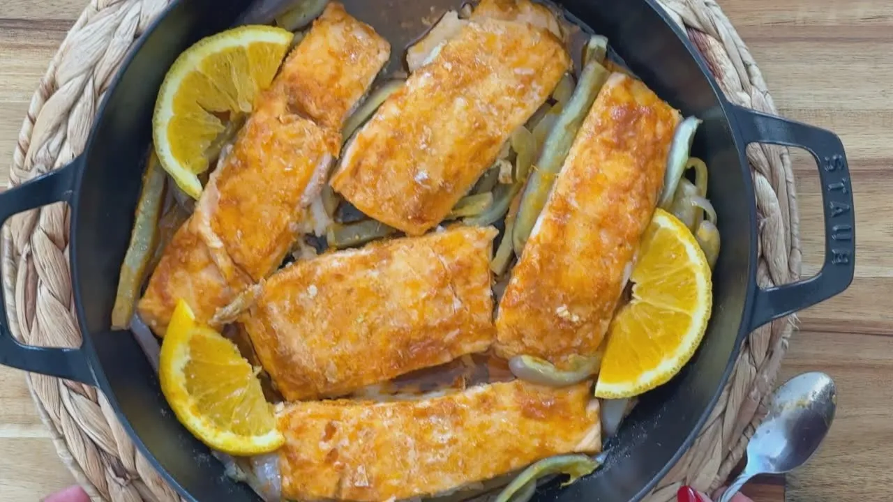 Baked salmon with oranges and vegetables in a black pan on woven mat, served on a wooden table.