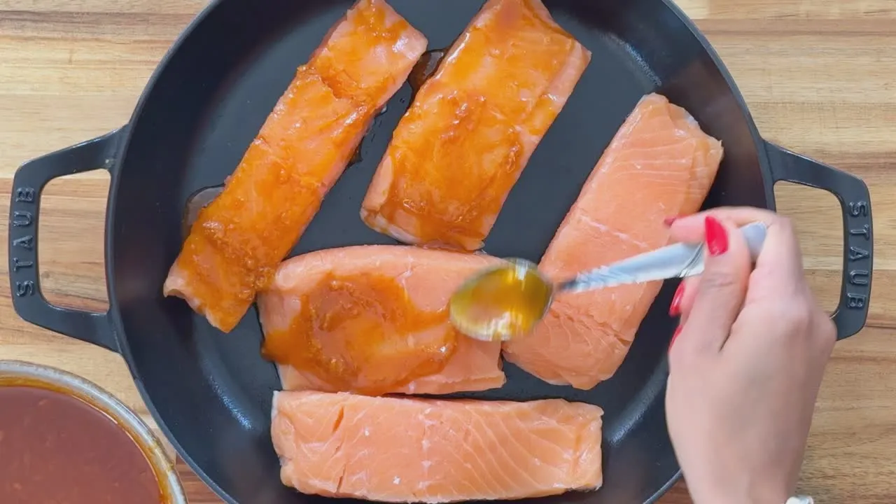 Person glazing salmon fillets with sauce in a pan on a wooden surface.