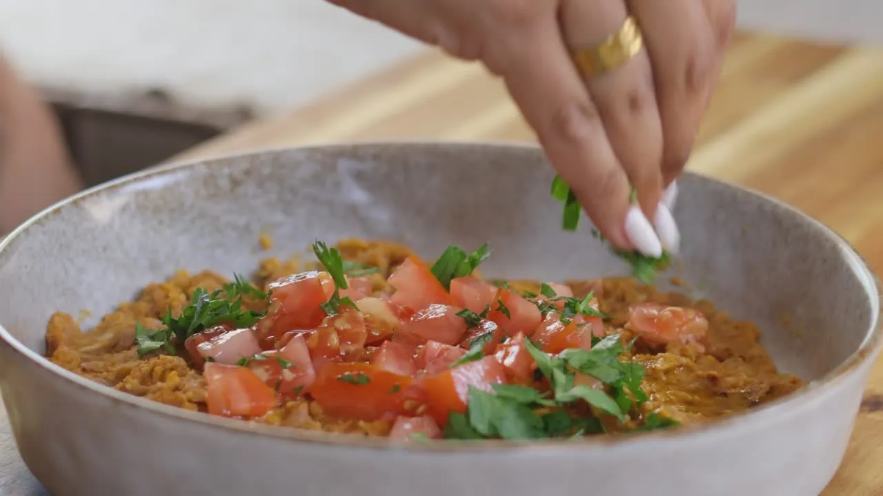 Hand garnishing dish with fresh tomatoes and herbs in a rustic bowl on wooden table.