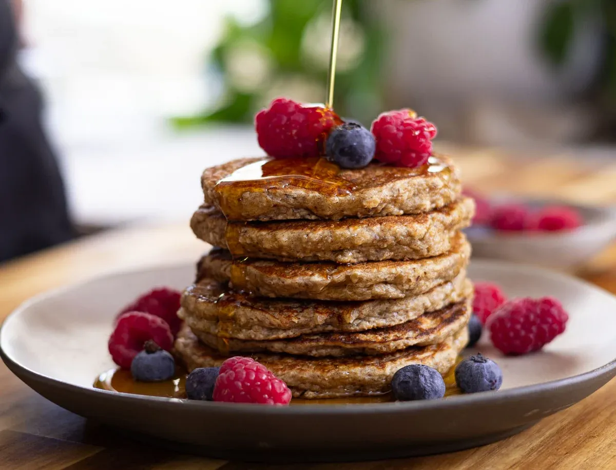 Stack of banana oatmeal pancakes topped with syrup, fresh raspberries, and blueberries on a plate.