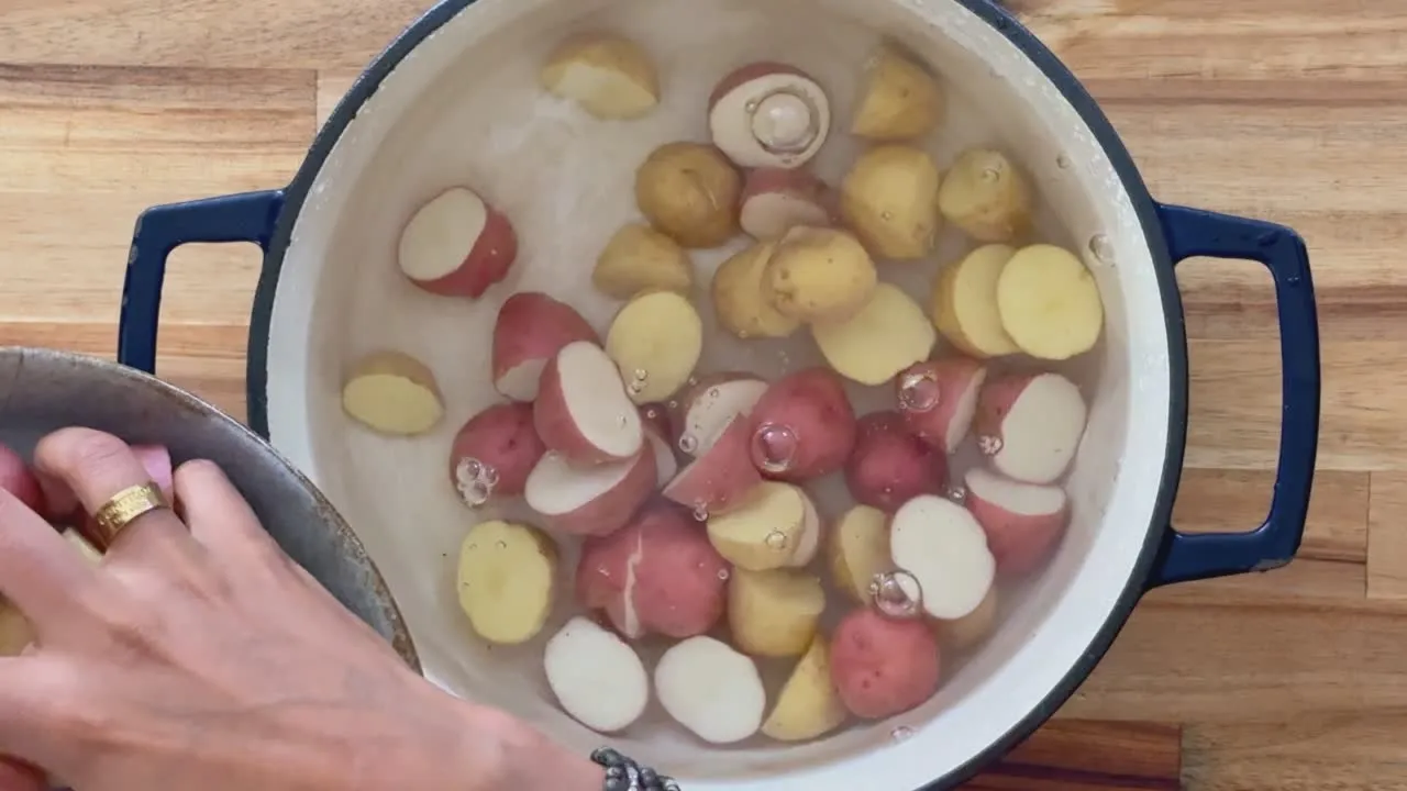 Person adding halved potatoes to pot of boiling water on wooden surface.