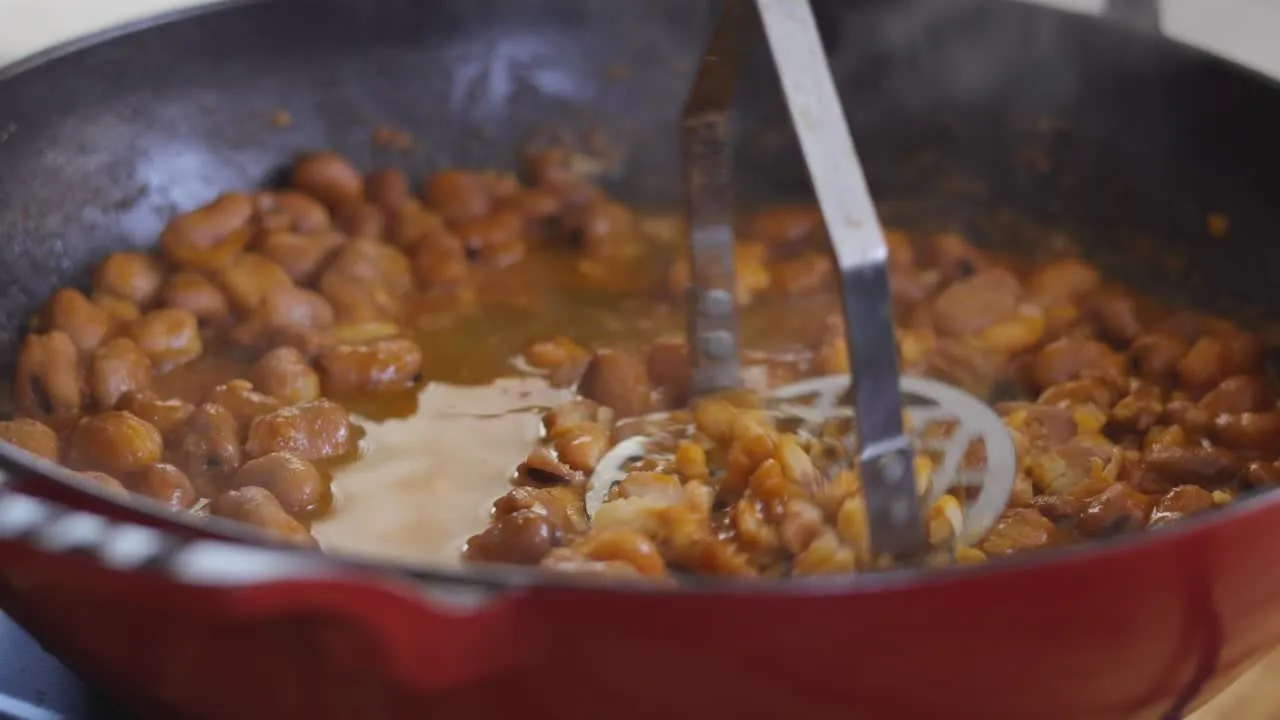 Mashed beans being prepared in a red skillet with a metal masher.
