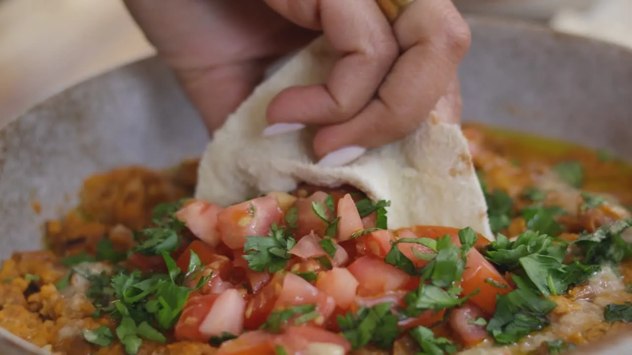 Hand dipping bread into savory dish topped with diced tomatoes and fresh herbs in a bowl.