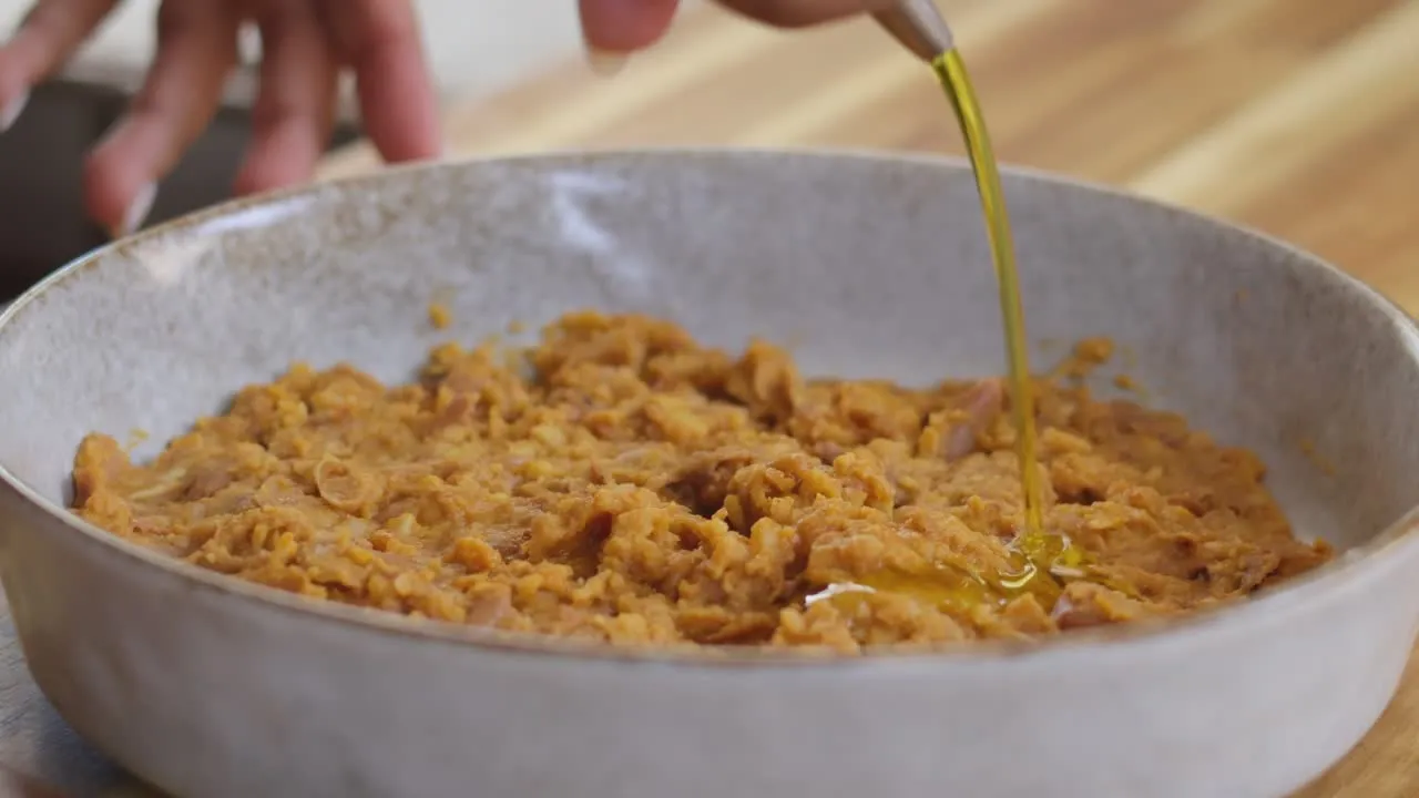 Pouring olive oil over mashed beans in a bowl on a wooden surface.