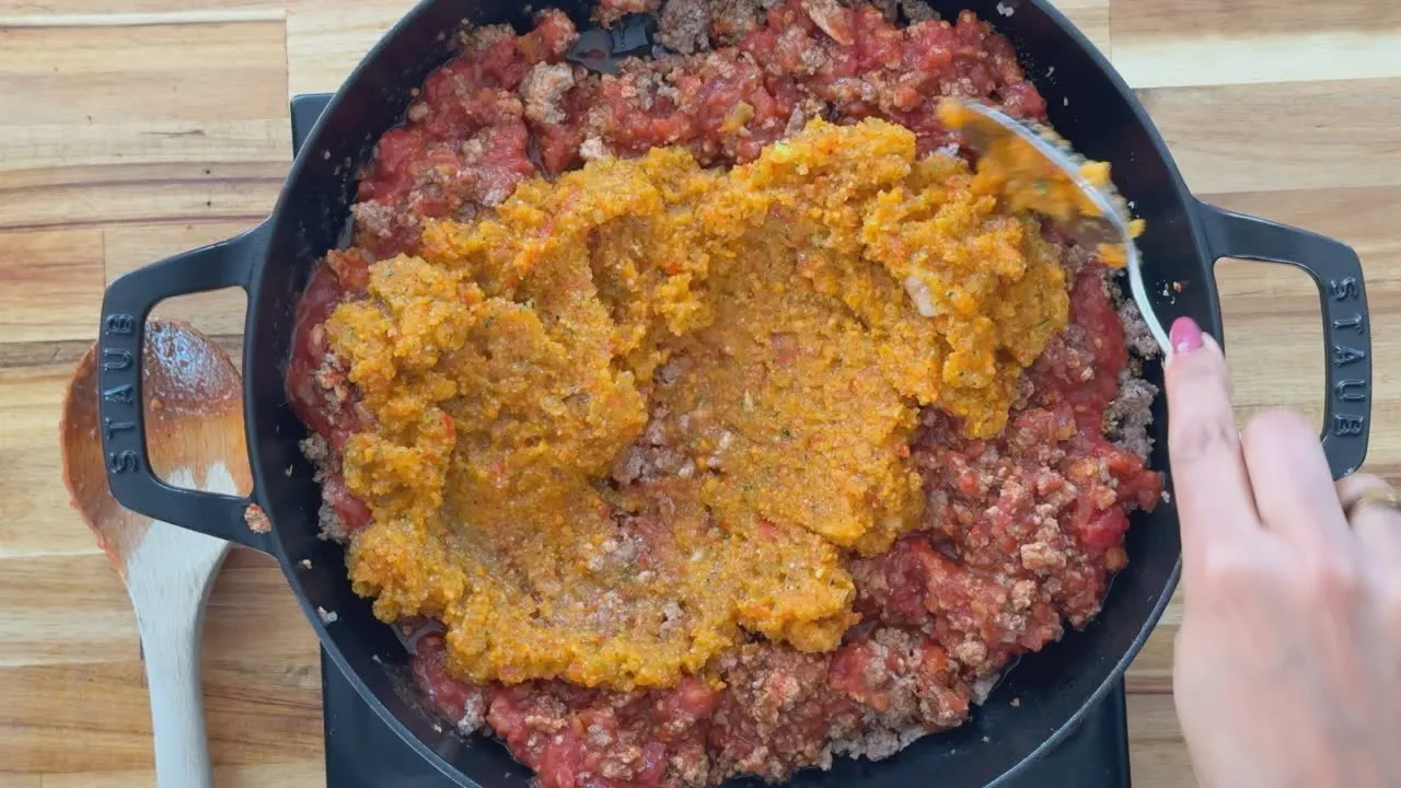 Cooking bolognese sauce in a Staub pan with beef, tomatoes, and vegetables being stirred by hand.