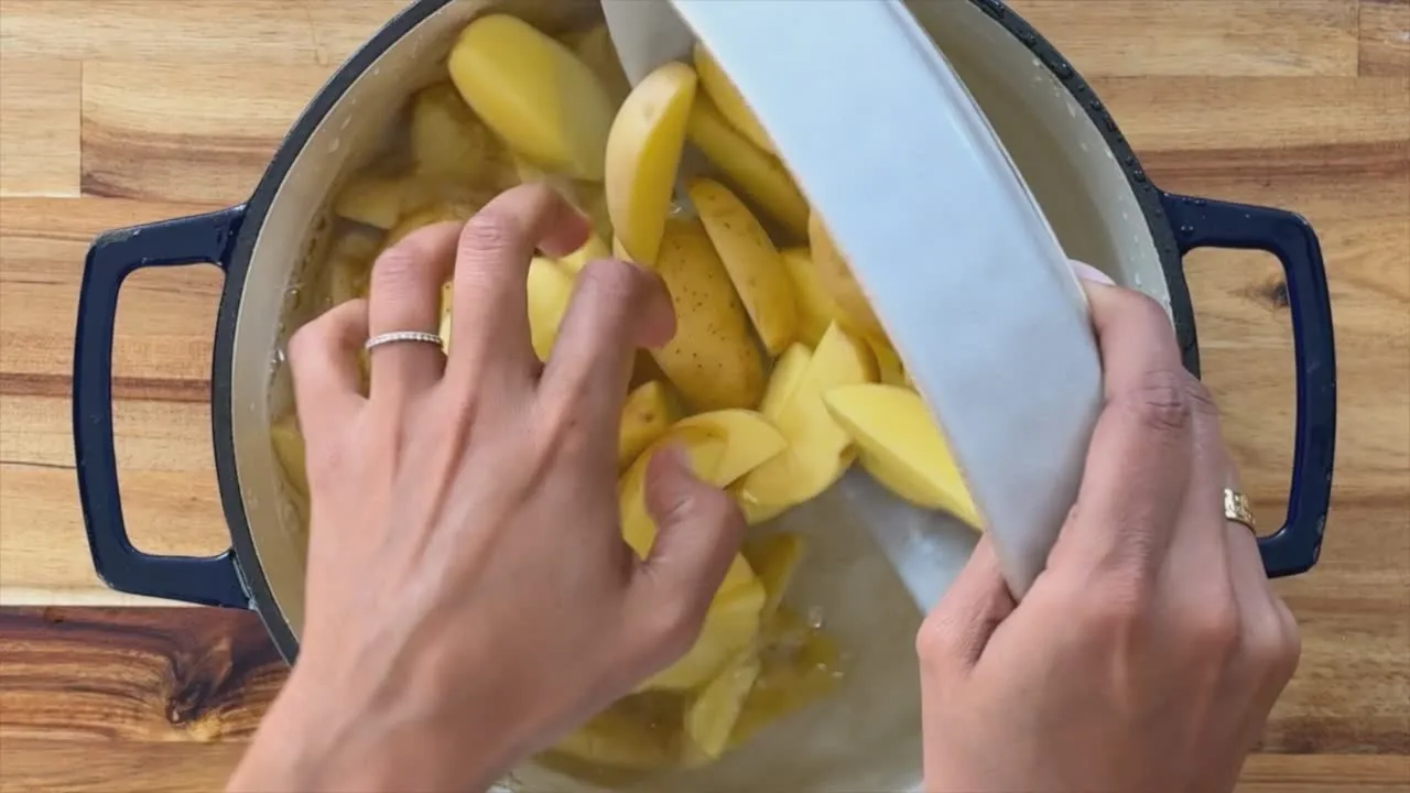 Person adding sliced potatoes into a pot on a wooden countertop.