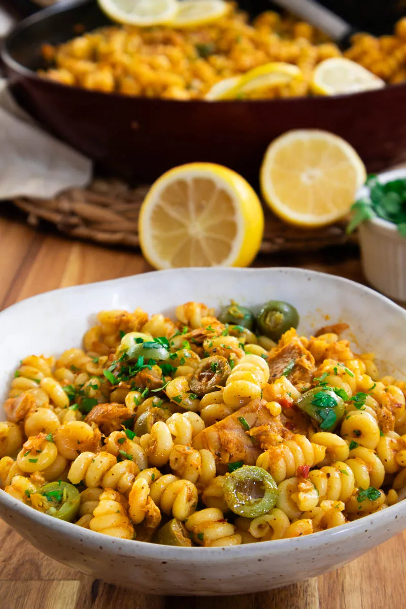 Bowl of spiral pasta with olives and herbs, garnished with lemon slices on a wooden table.