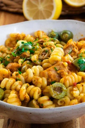 Bowl of Creamy Calabrian Tuna Pasta with green olives, parsley, and chicken on a wooden table. Lemon slice in the background.