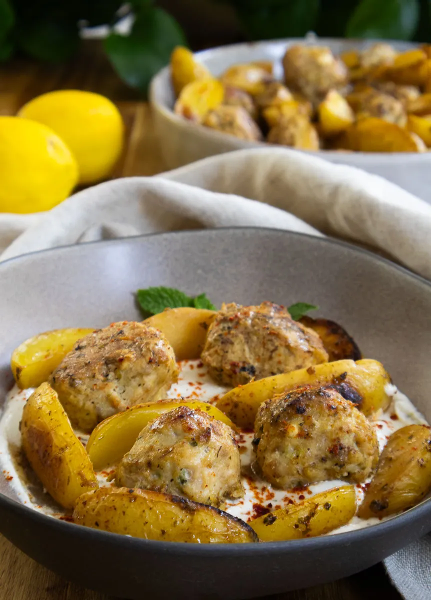 Bowl of Greek Chicken Feta meatballs with roasted potatoes and whipped feta, garnished with herbs and lemons.
