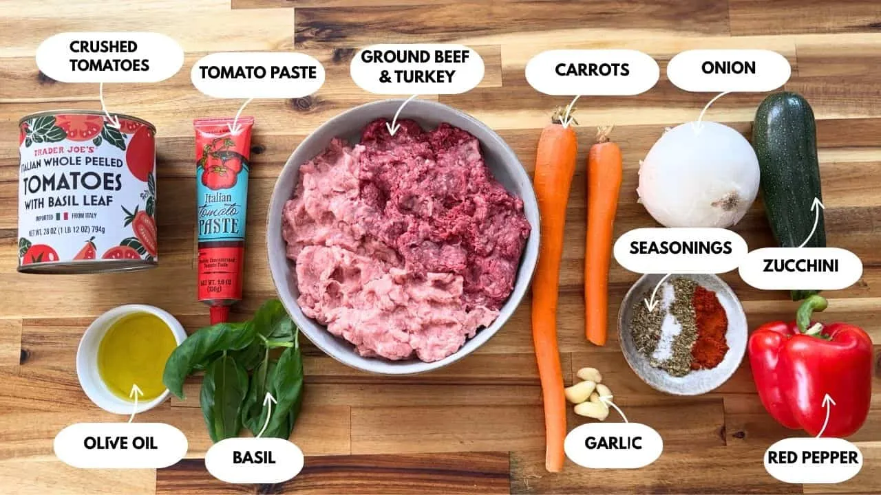 The ingredients for homemade hidden veggie pasta sauce are ground beef, tomatoes, paste, veggies, spices, basil, and olive oil, which are served on a wooden table.