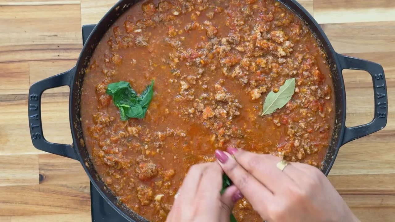 Hands adding fresh herbs to simmering tomato meat sauce in a Staub pot on a wooden surface.
