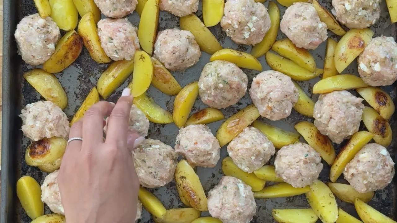 Hand arranging meatballs and potato wedges on a baking tray for roasting.