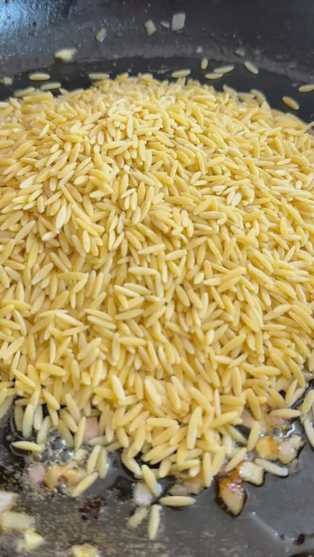 Close-up of dry orzo pasta cooking in a frying pan with some chopped garlic and onions.