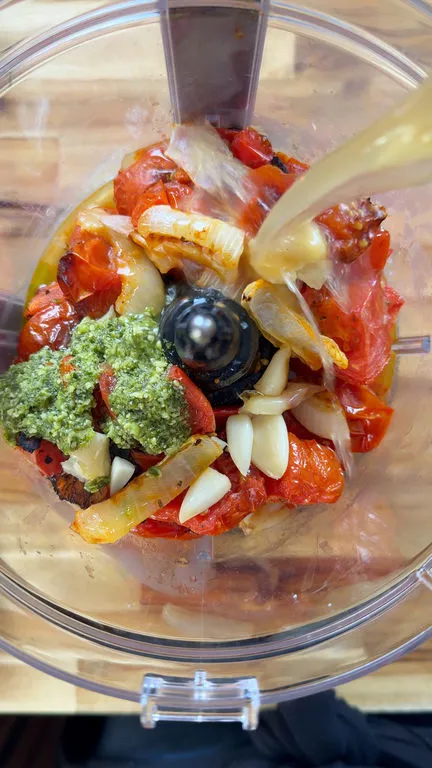 Food processor with tomatoes, pesto, garlic, and onions being blended from above.