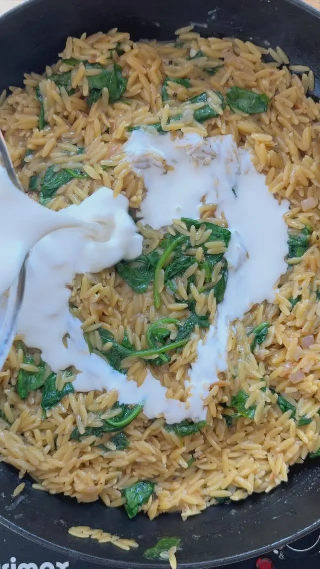Creamy orzo pasta with fresh spinach being mixed in a pan drizzled with yogurt.