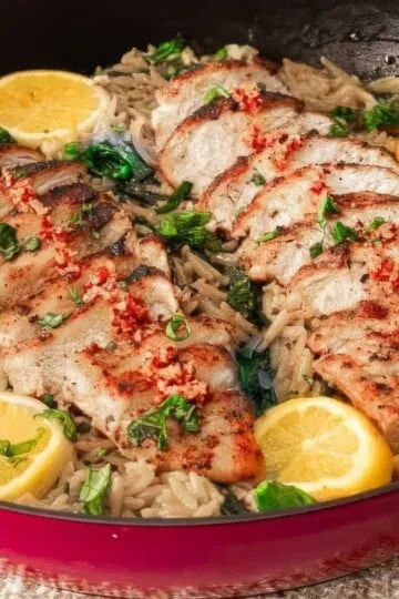 Grilled chicken with herbs on rice in a skillet, garnished with lemon slices and fresh greens.