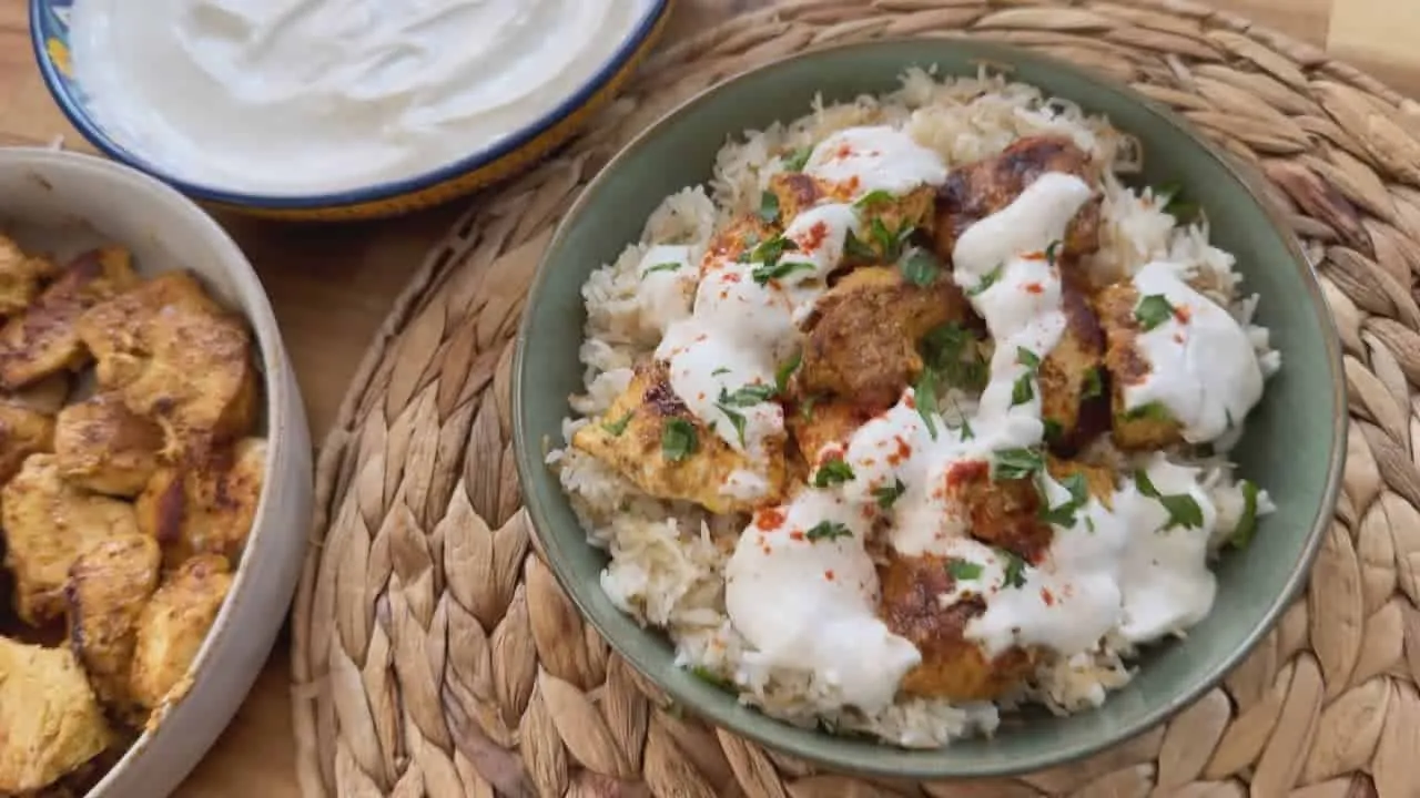 Bowl of spiced chicken over rice topped with yogurt sauce, next to a bowl of plain chicken, on a woven mat.