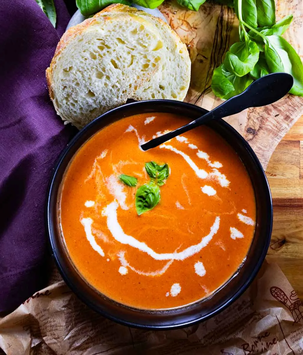 Roasted cherry & heirloom tomato soup garnished with basil leaves, served with rustic bread on a wooden table with a purple cloth.