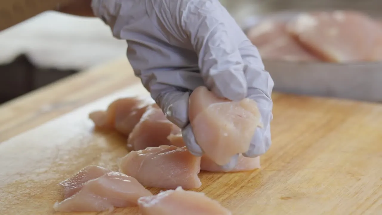 Gloved hand handling raw chicken pieces on a wooden board for cooking preparation.