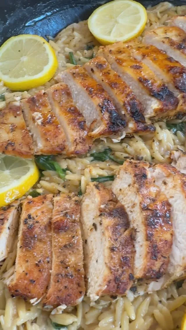 Grilled chicken slices over creamy orzo pasta topped with lemon slices in a skillet.