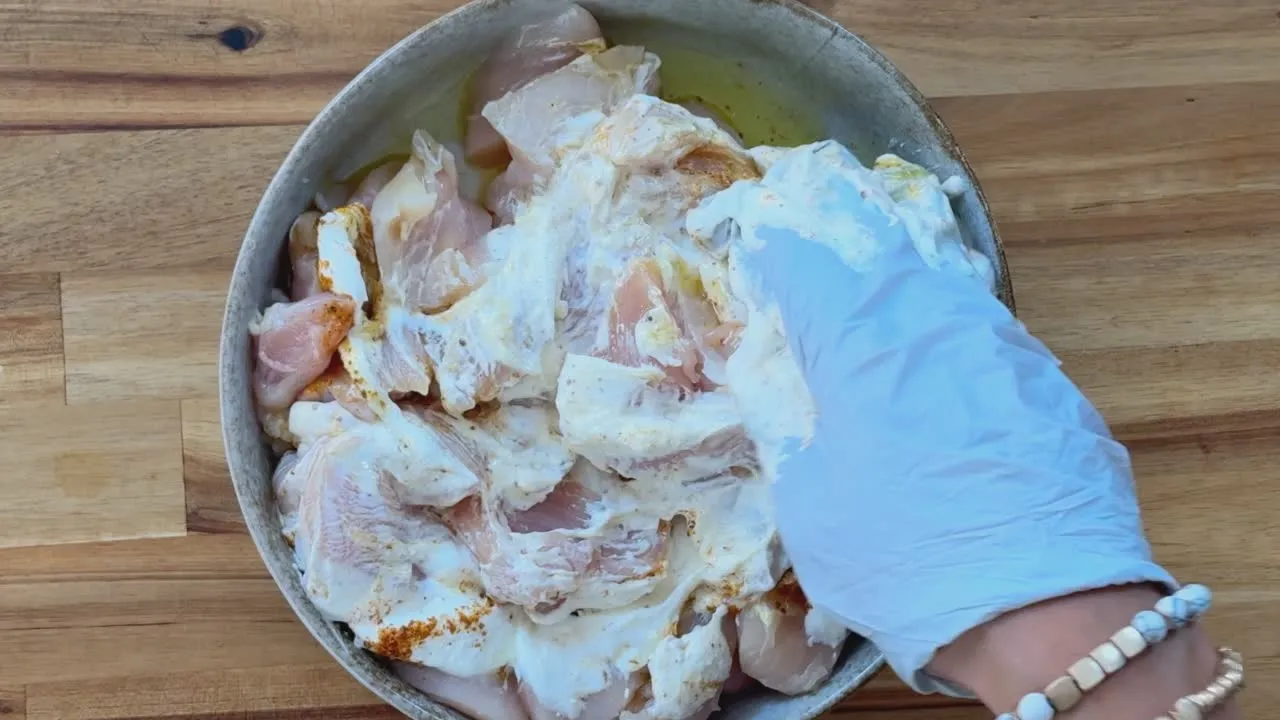 Marinating chicken pieces with yogurt and spices in a bowl, using a gloved hand for mixing on a wooden surface.