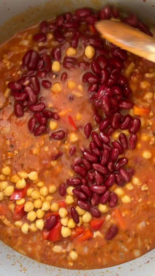 Simmering stew with red kidney beans, chickpeas, and chopped red peppers being stirred with a wooden spoon.