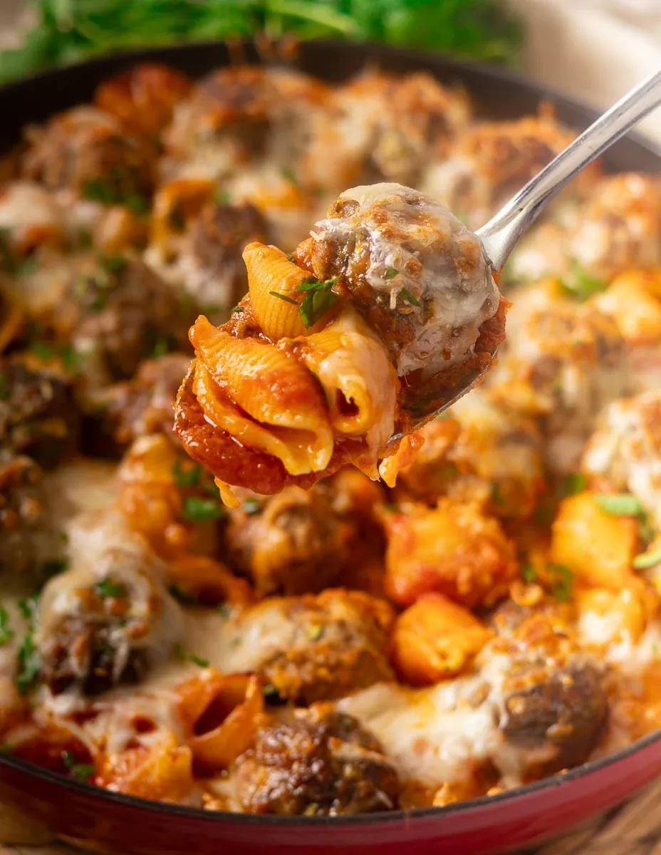 Cheesy baked meatballs and pasta with tomato sauce on a spoon over a skillet, garnished with herbs.