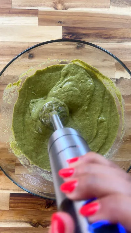 Blending vegetable soup in a glass bowl with a hand blender on a wooden surface.