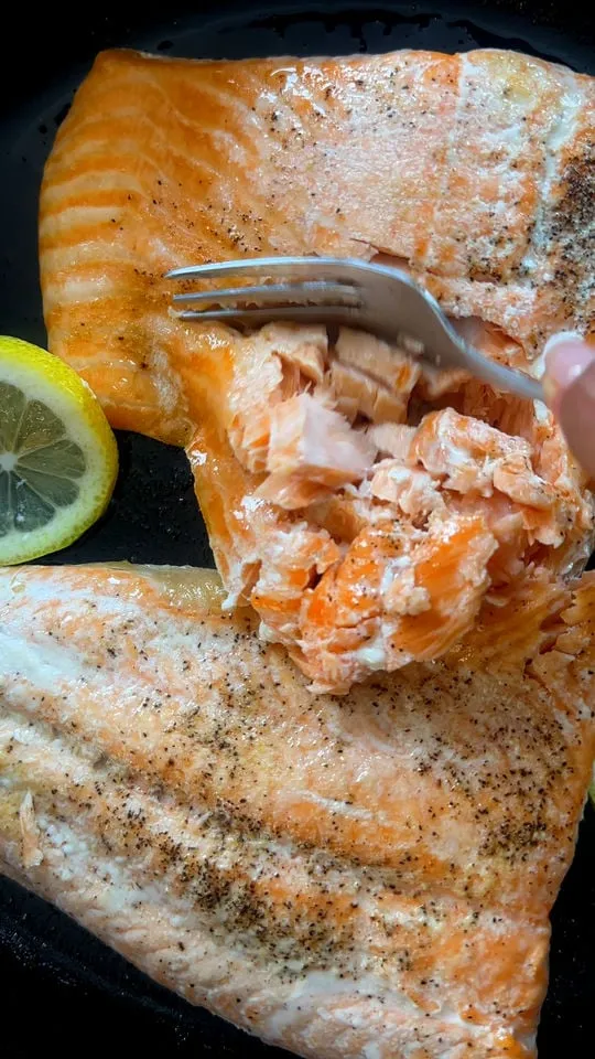 Flaky cooked salmon on a skillet with lemon slice and fork, seasoned with pepper.