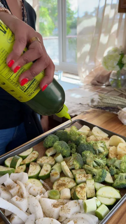 Person drizzling olive oil on seasoned vegetables in a tray, preparing for roasting.