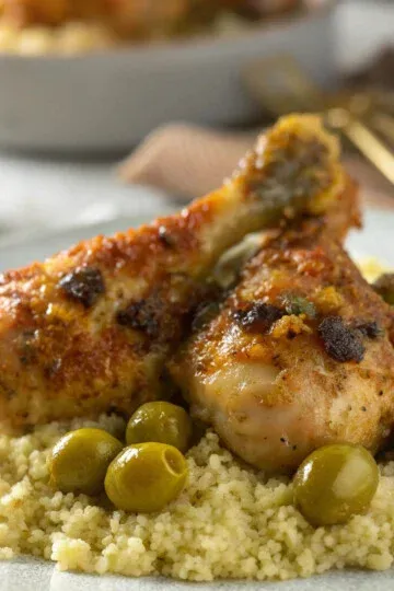 Moroccan lemon chicken and olives served on fluffy couscous, garnished with herbs, on a white plate.