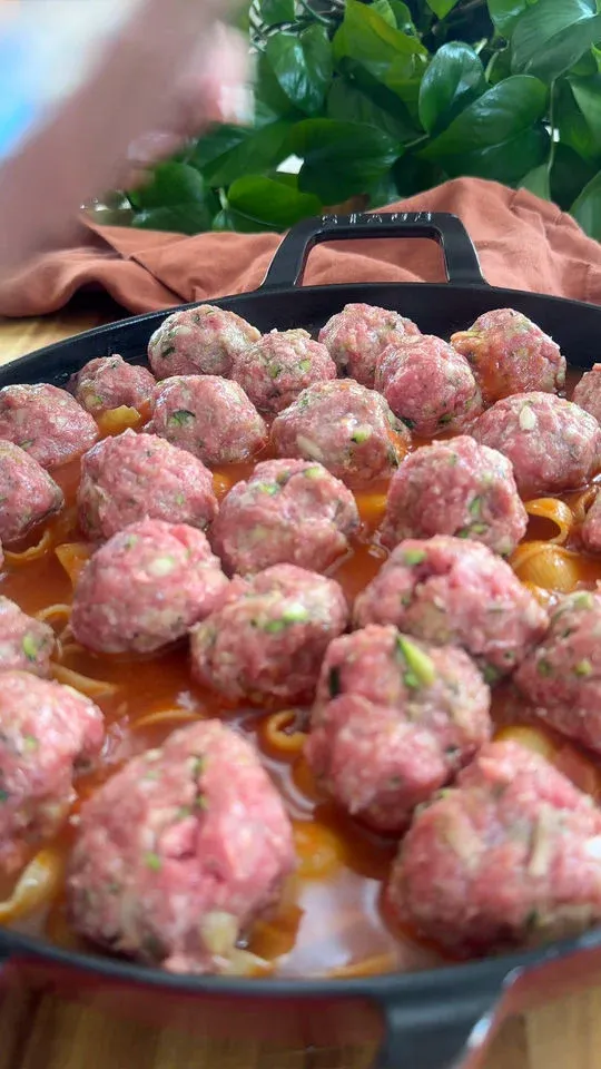 Raw meatballs in tomato sauce in a black pan with a plant and brown cloth in the background.