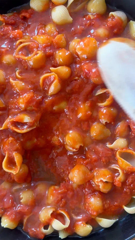 Shell pasta mixed with chunky tomato sauce and a wooden spoon in a pan.
