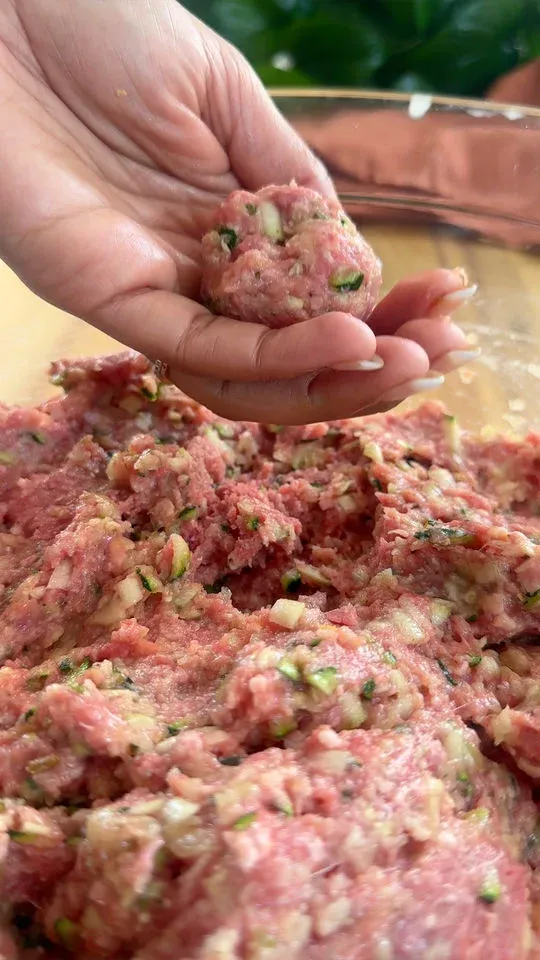 Hand shaping meatball with minced meat mixture in a glass bowl.