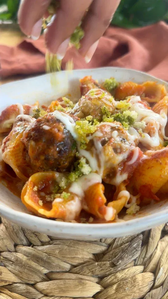 Hand sprinkling herbs on pasta with meatballs and melted cheese in a white bowl.