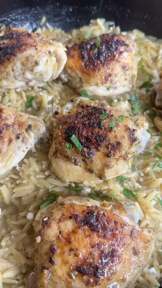 Close-up of browned chicken thighs with orzo pasta, garnished with fresh herbs in a skillet.