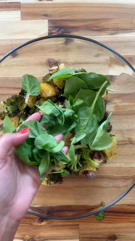 Hand adding fresh spinach to a bowl of roasted vegetables on a wooden table.