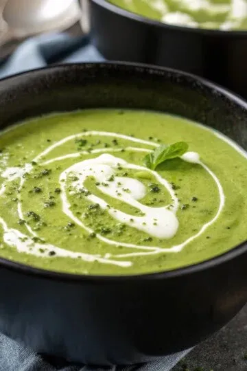 Creamy green soup garnished with herbs and a swirl of cream in a black bowl on a wooden table.